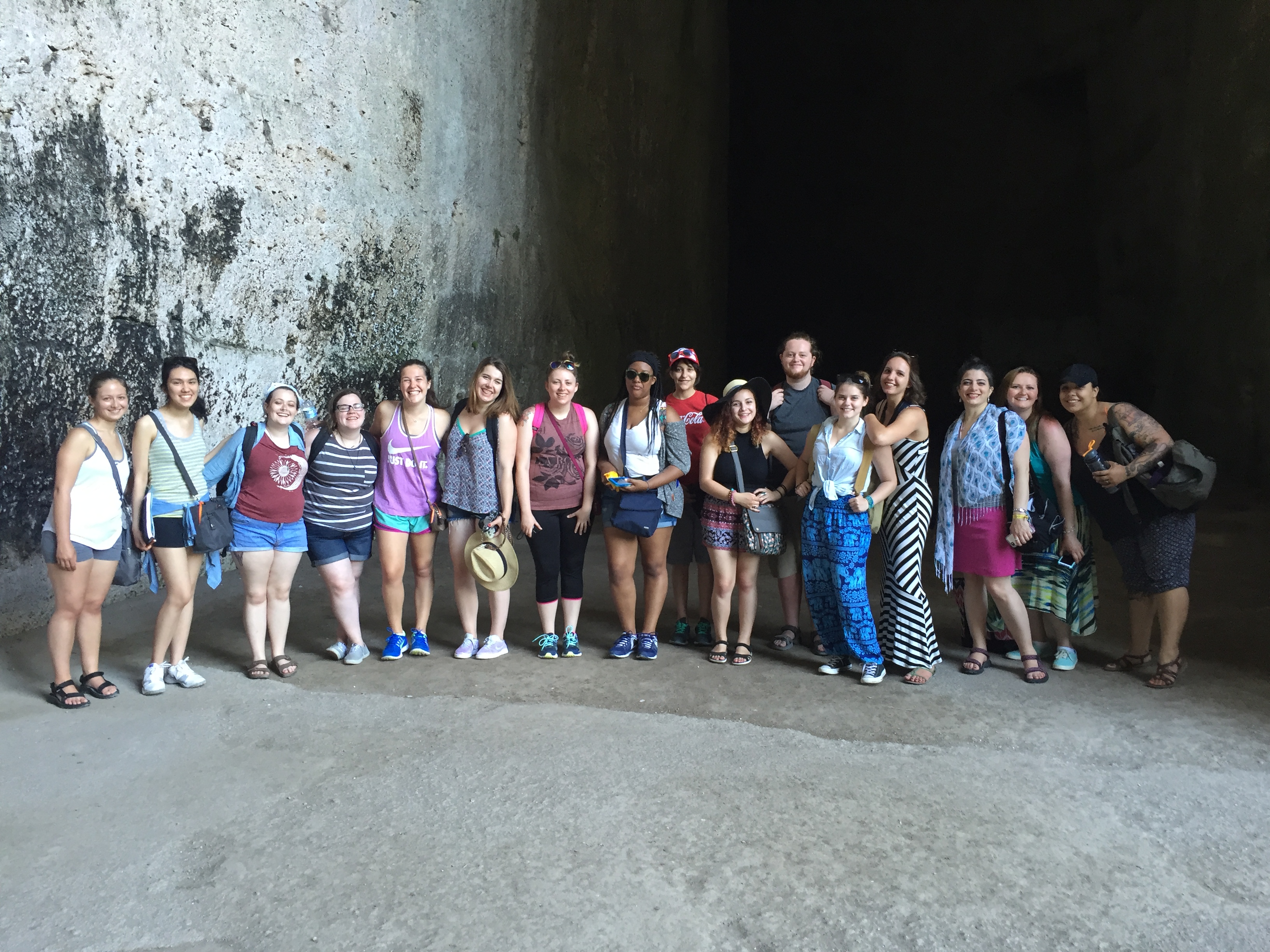 Group photo of 16 students from On-Site Studies in Art History Abroad Summer 2016 program, The Greeks in Ancient Italy. The students are posed at the mouth of an ancient rock quarry cave site in Syracuse, known as the 'Ear of Dionysios.' The cave is known for its exceptional acoustics: noises echo, but repeat only once. Ancient tyrants eavesdropped on prisoners they held captive here.
